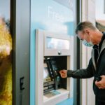 A man wearing a mask using an ATM machine outdoors for cash withdrawal.